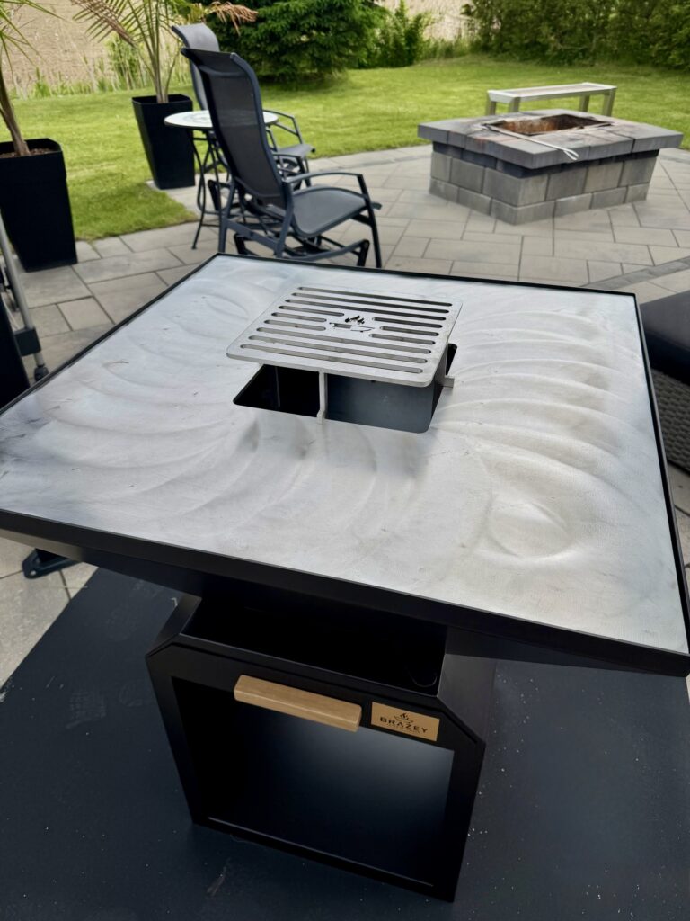 Close-up of a stainless steel grill vent on a black outdoor grill on a paved patio with chairs and a fire pit nearby.