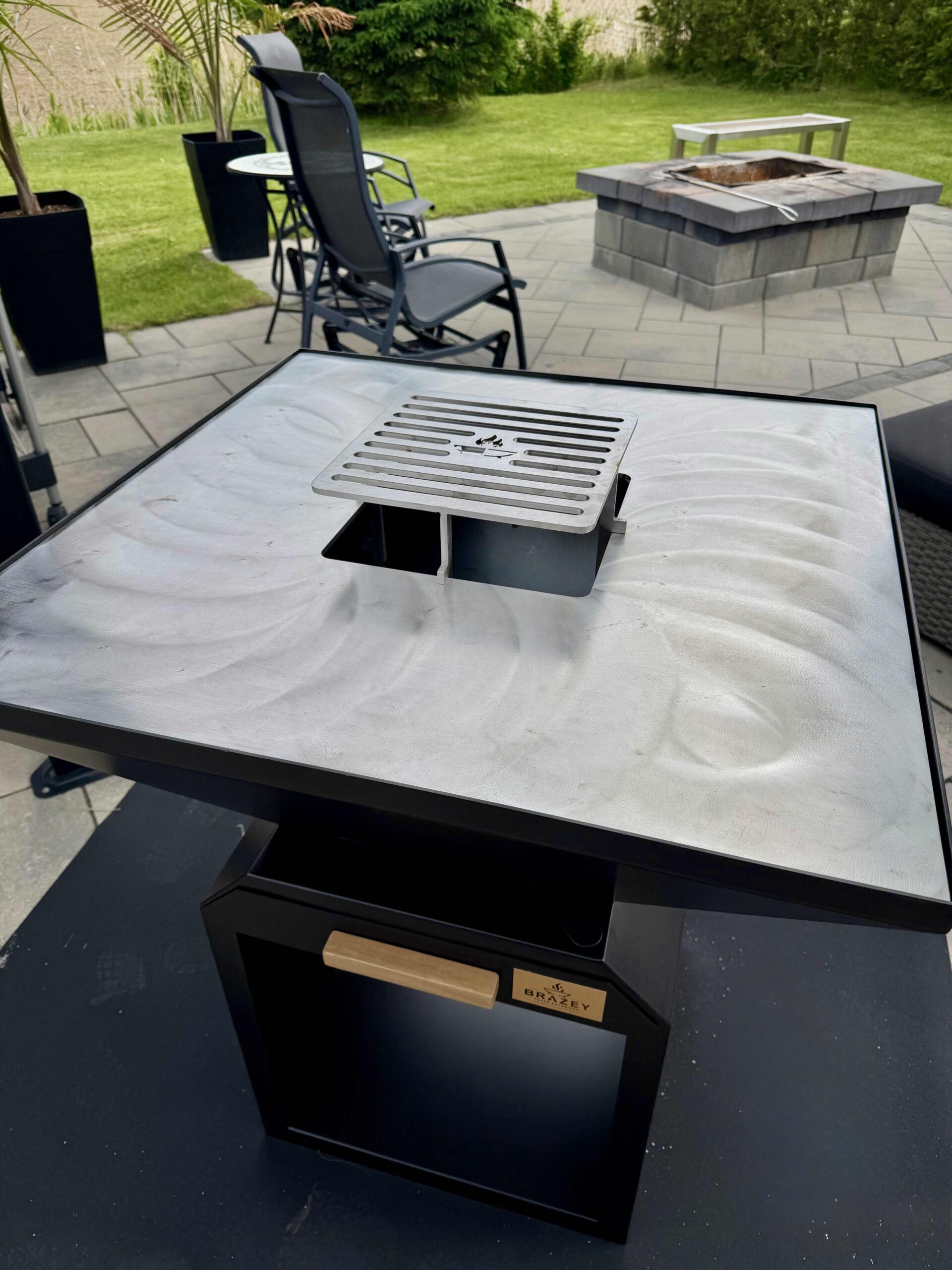 Stainless steel grill grate mounted on a glass outdoor table, with a brick fire pit and patio chairs in the background.