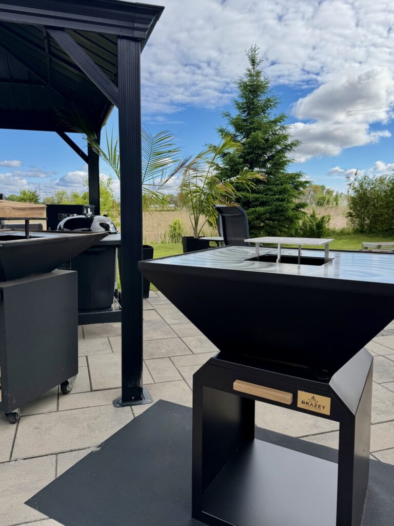 Outdoor black metal barbecue grill area with a large modern grill and prep table, under a dark pergola, with trees and blue sky in the background